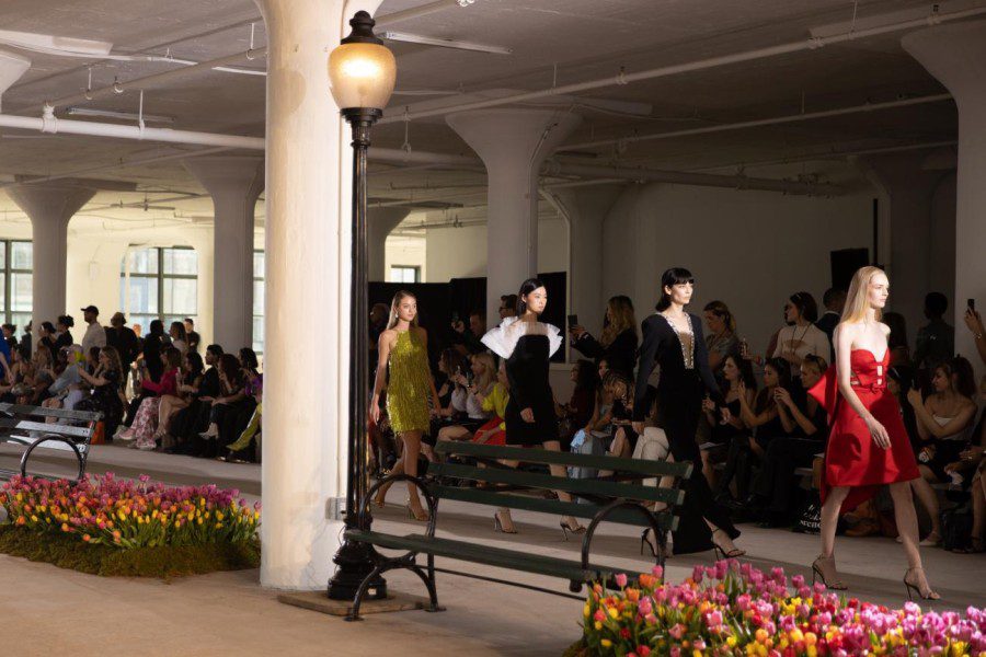 A group of models walk down the runway, one in a green dress, one in a black-and-white dress, one in a black dress and one in a red dress.