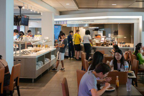 Students walk and dine in an indoor space with dining tables and chairs in the foreground and there are food court vendors in the background.