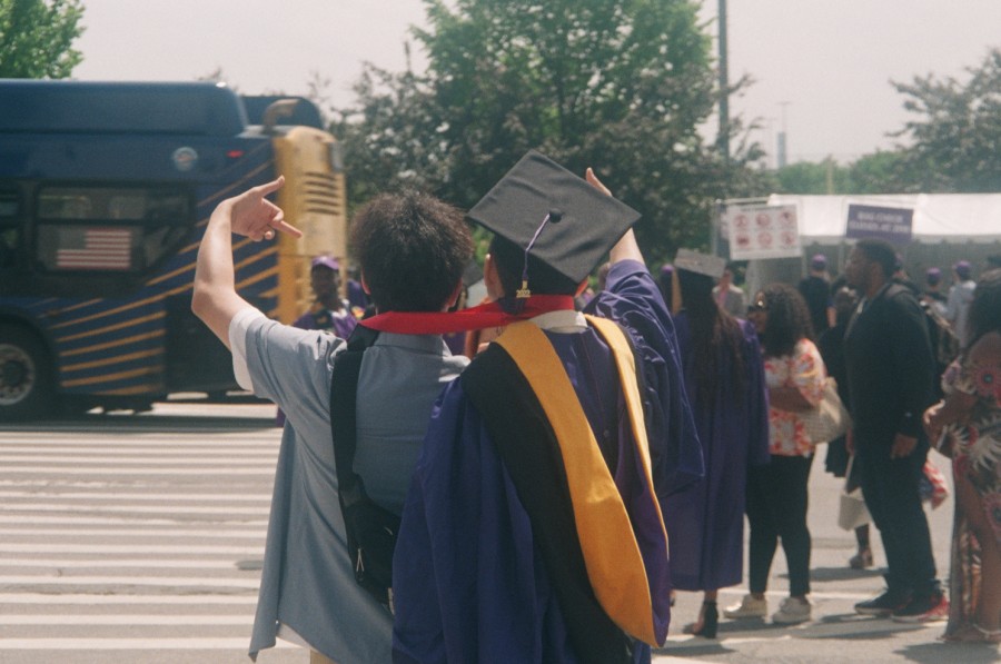 A person in a gray shirt stands to the left of a student at a graduation ceremony. They are posing in front of a crowd of graduates with their families and a moving bus.
