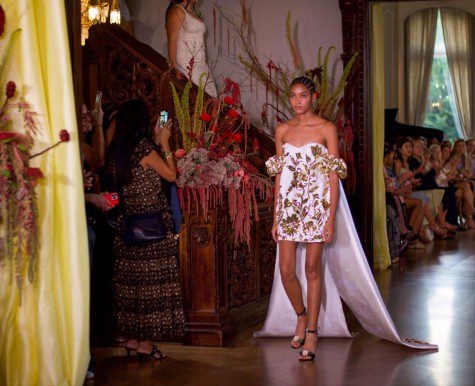 A model walks down the runway in an off-the-shoulder white mini dress with metallic gold floral embellishments. Two long cape-like trains are attached to its back.