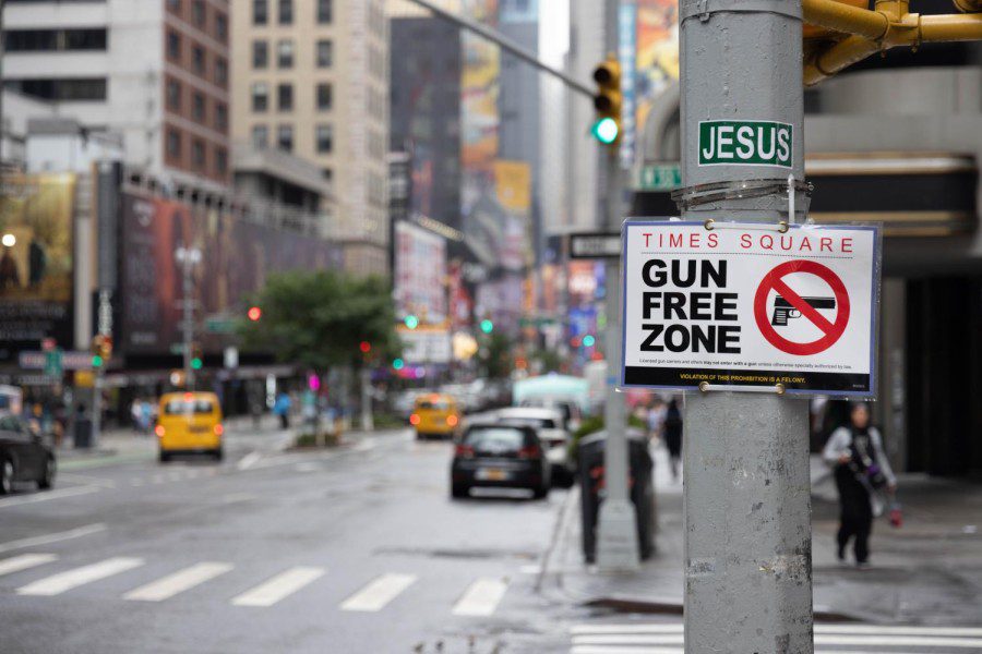In the foreground is a pole with a sign reading “Times Square: Gun Free Zone” and the 53rd Street streetscape in the background.