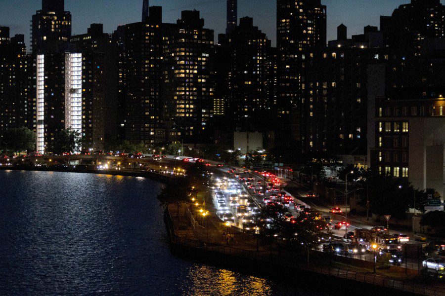 A riverside view of Manhattan at night. There is two-way traffic on a crowded highway with high rises in the background.