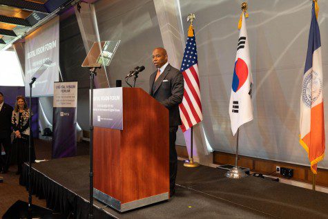 A man wearing a suit stands behind a wooden podium and stands on a black stage. Behind him there are three flags: the red, white and blue U.S. flag; the Korean red, blue, white and black flag; and an orange, white and blue flag.