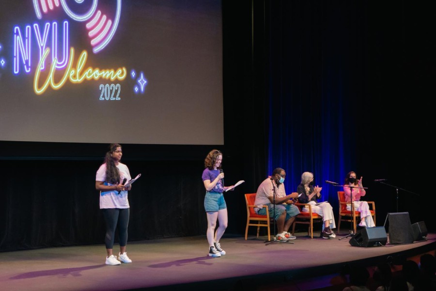 A stage with “NYU Welcome” projected on the screen. Two speakers standing on the right and three guests sitting on the right.