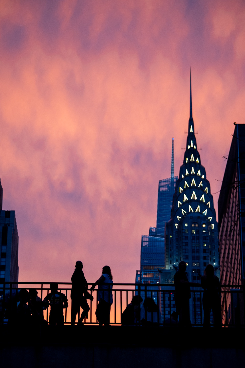 A group of people standing on an overpass. It is just after sunset, and the sky is lit purple. The Chrysler building is in the background.