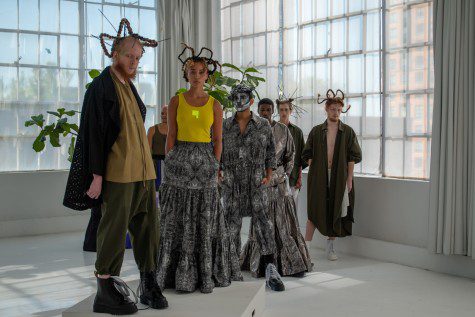 Seven models stand on a platform dressed in various oversized garments. All wear devil-like horns.