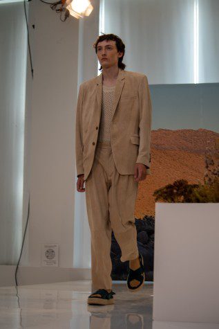 A male model walks in a tan suit with broad shoulders against a desert image as background.