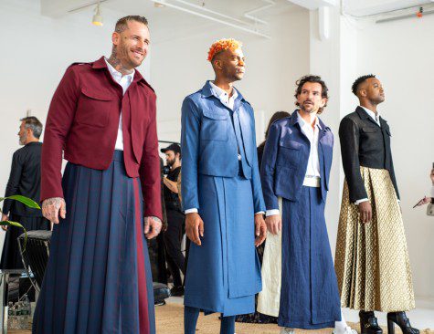 Four male models stand while dressed in various jackets and full-length skirts.