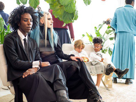 A male model wearing a black suit, white shirt and black tie sits at the left; a pale-faced female model dressed like a medieval nun sits to the right of the male model. Two children sit at the right.