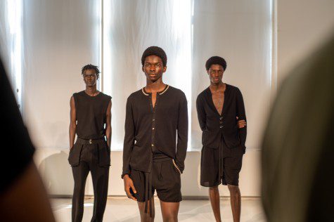 Three male models stand in front of a white screen dressed in black cardigans and shorts.