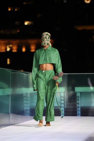 Model walking down aisle in a green two-piece pantsuit with an emerald and gold mask.