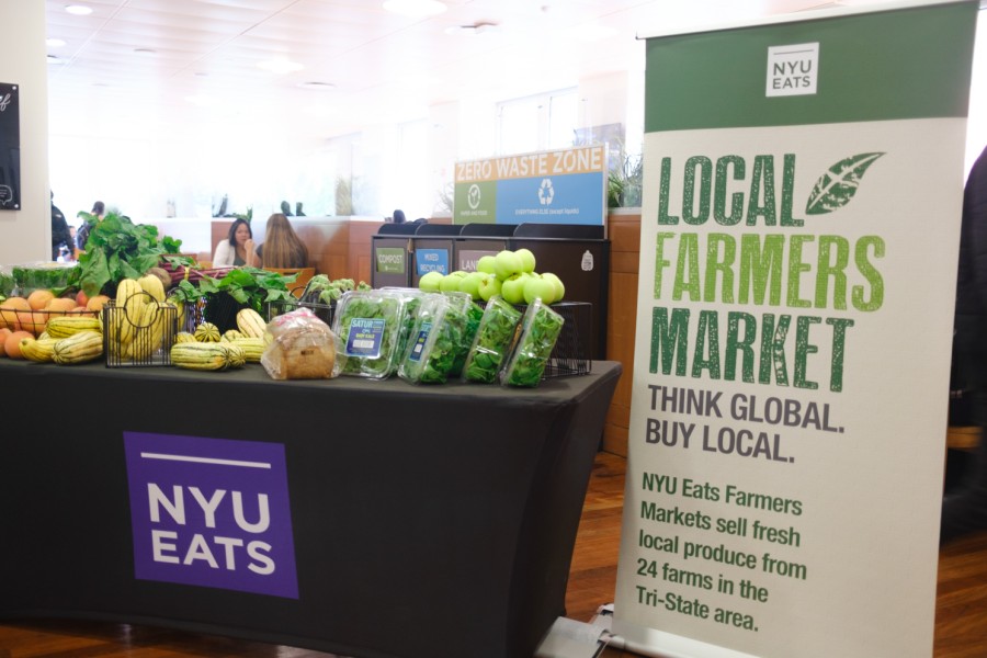 Photograph of a banner saying “NYU Farmer’s Market” on the right, and an assortment of fruits and vegetables on a table covered in a purple NYU-branded tablecloth on the left.