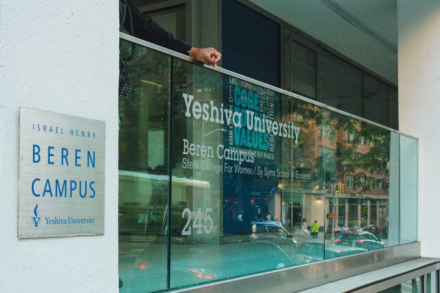 A plaque says “Israel Henry Beren Campus, and under the text a logo of a torch next to the words Yeshiva University.” The plaque is plastered next to a glass balcony of Yeshiva University’s Stern College for Women. A person stands behind the balcony.