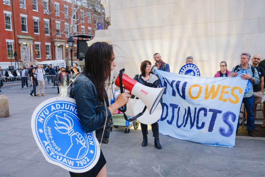 A speaker with a red megaphone carrying a blue banner that reads “N.Y.U Adjunct Union A.C.T-U.A.W Local 7902” shouts into a crowd of union workers under the Washington Square Arch. There are people behind the speaker holding other signs.