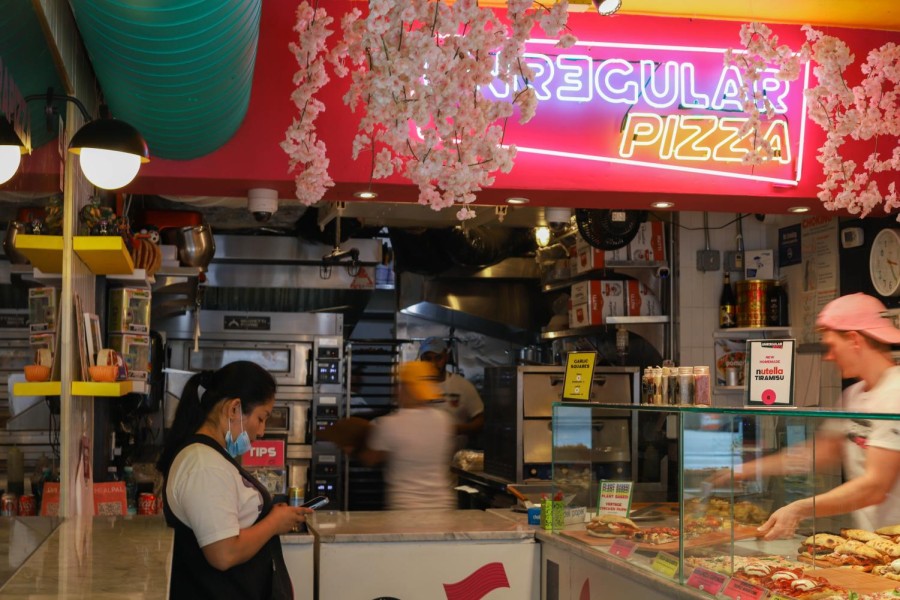 The interior view of Unregular Pizza. There are one customer and three chefs in the frame. Purple Neon sign saying “Unregular Pizza” hangs above.