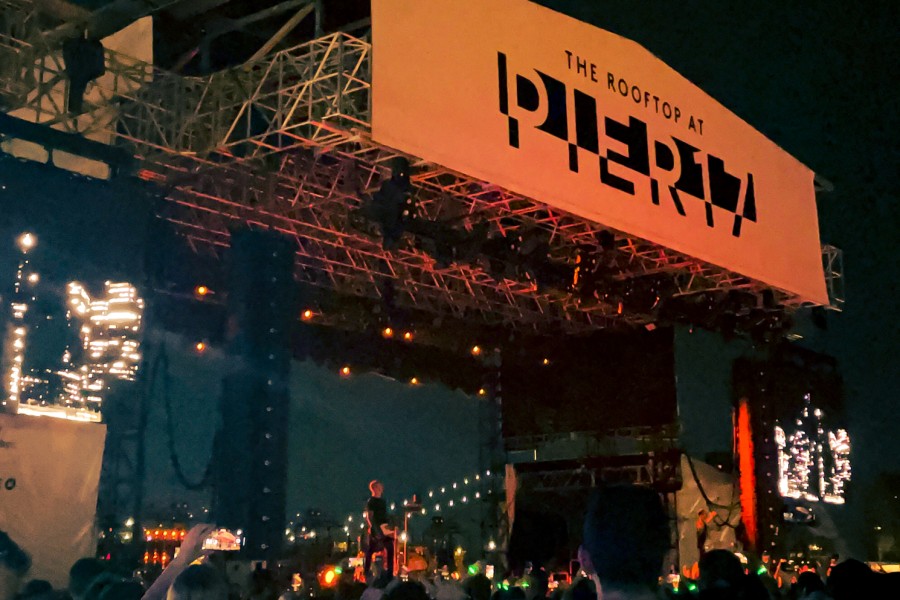 A crowd of concert attendees stands in the foreground with a stage and a male performer singing on stage in the background.