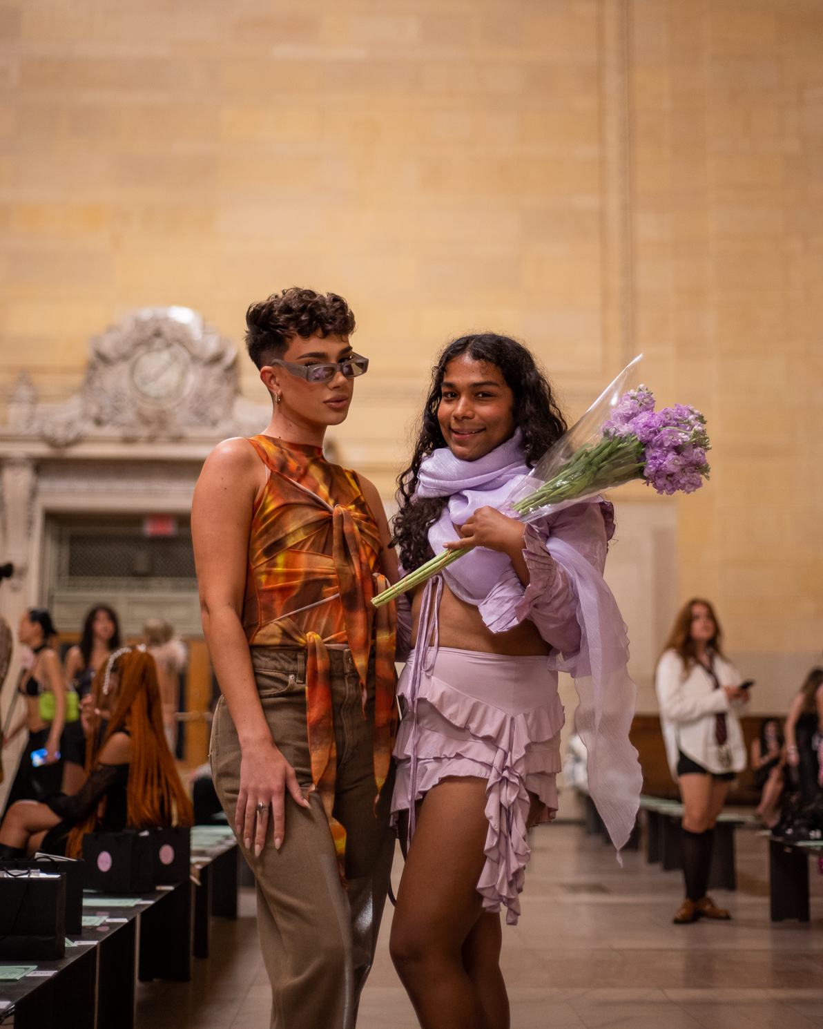 James Charles wearing an orange printed top and Devin Halbal wearing a purple outfit stand in Grand Central Terminal.