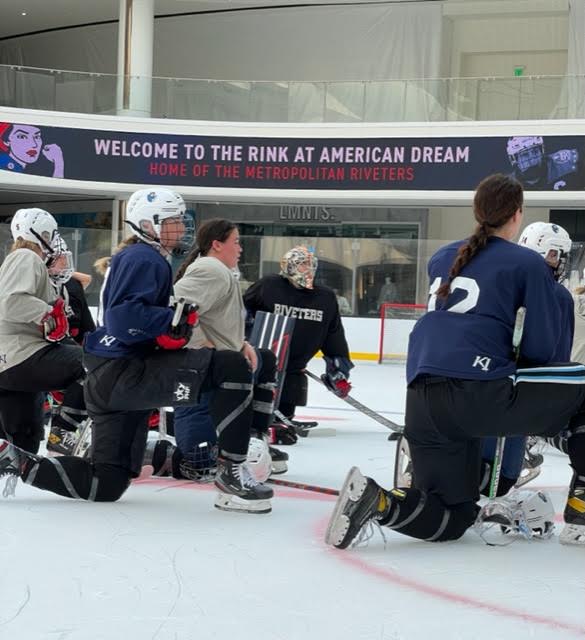 A group of Riveters players kneel inside of the American Dream mall ice rink, wearing sports jerseys and ice hockey gear.