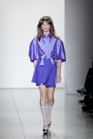 A female model wearing white stockings and heels walks in a reflective purple blouse with a ribbon at the waist and a dark purple skirt.
