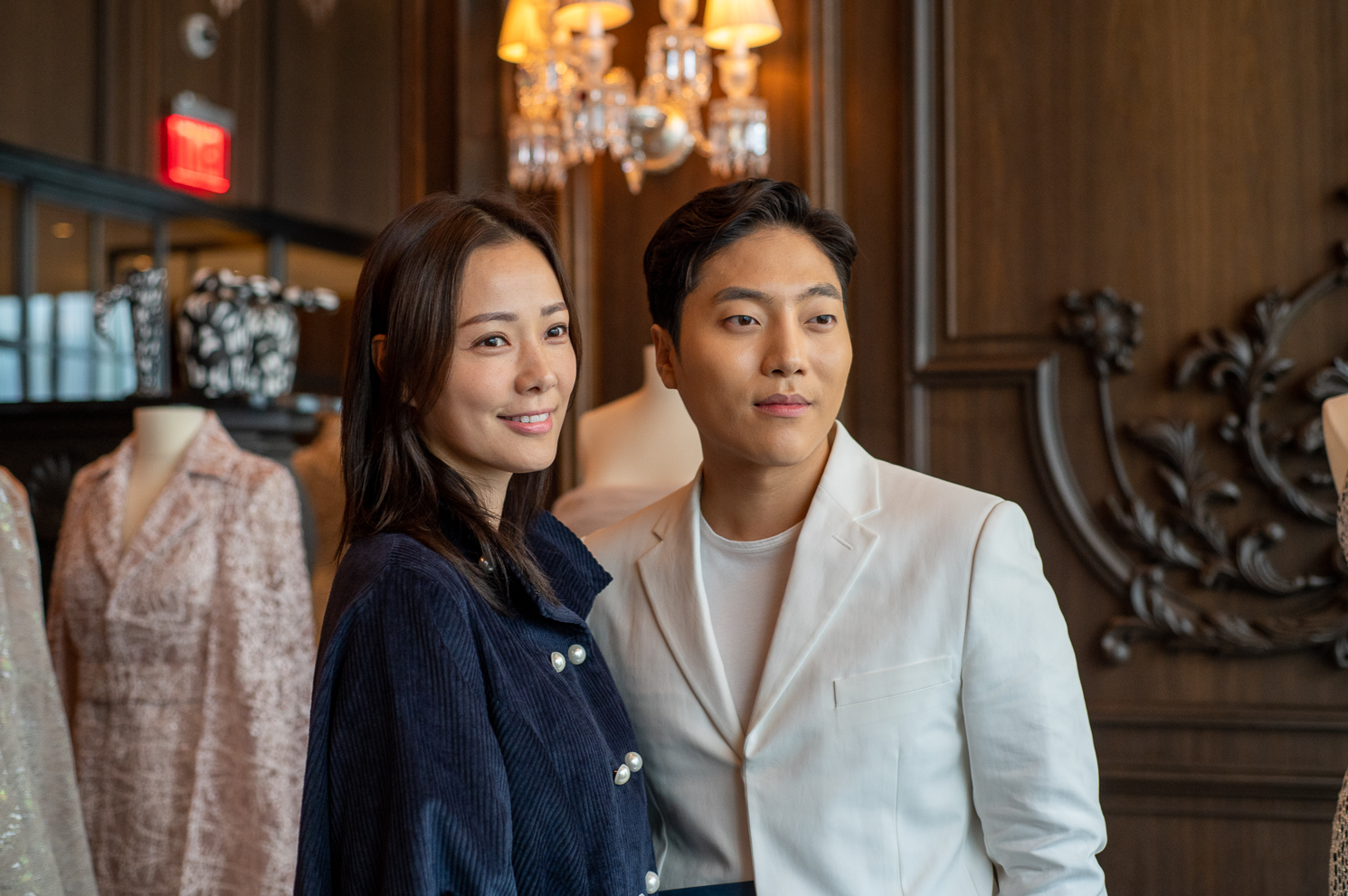 Son Tae-young wearing a navy jacket and Andrew Kwon wearing a white suit, standing in front of a chandelier.