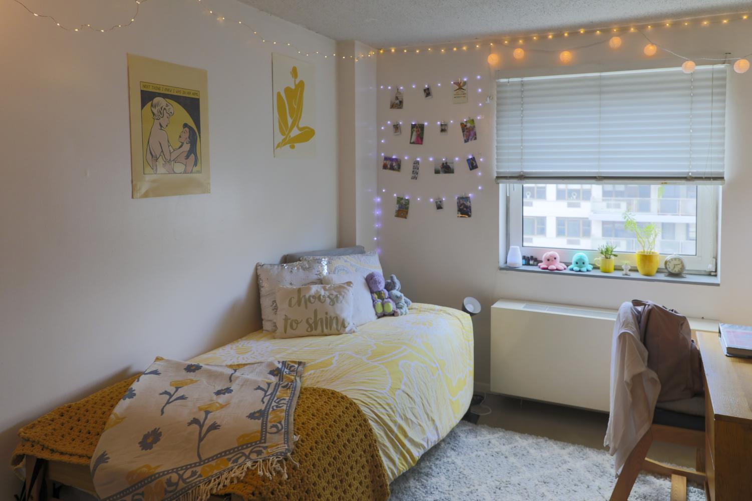 Lily Candelaria's dorm room has her bed on the left side with yellow accents and decorative pillows on top. The wall above the bed features two decorative posters and a string of Polaroid photos of Lily’s friends and family. Lily’s windowsill has many decorative pieces sitting on it.