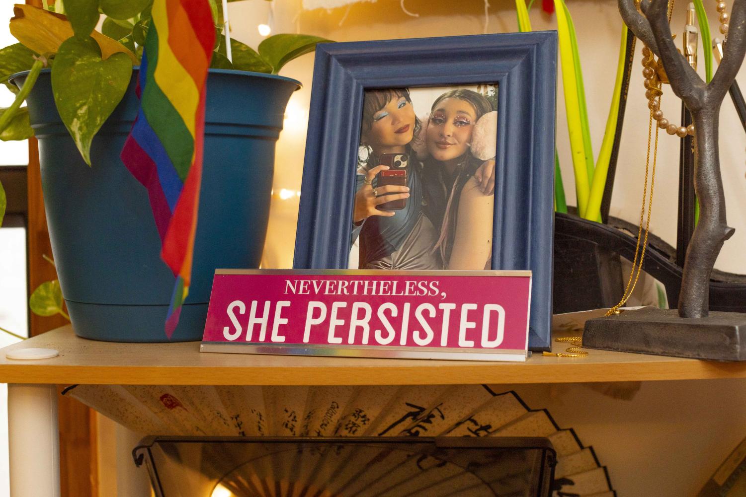 On top of Natalie Choi’s bedroom shelving is a blue planter with a small plant, a LGBTQ+ pride flag, a photo of Natalie and her friend, a small sign that reads “She Persisted” and a tree-like jewelry holder.