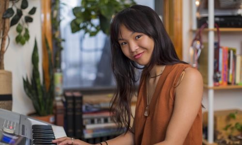 Natalie Choi looks at the camera while sitting down and playing her piano. Natalie has a smiling expression. Natalie is wearing a rust-colored blouse.