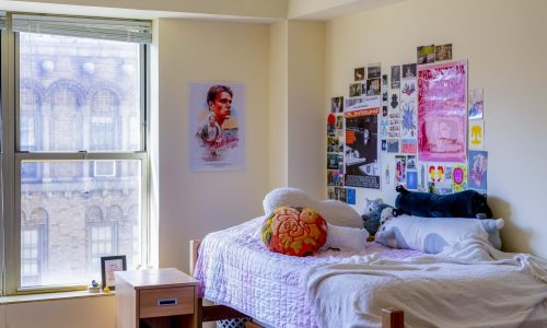 The interior of Lizzie England's dorm room at Clark Hall. Next to a window is Lizzie's bed, a poster of the movie “Good Will Hunting” and various collages of pictures. On Lizzie's bed lie various pillows and stuffed animals.