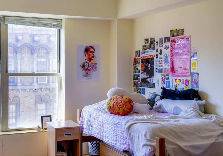The interior of Lizzie Englands dorm room at Clark Hall. Next to a window is Lizzies bed, a poster of the movie “Good Will Hunting” and various collages of pictures. On Lizzies bed lie various pillows and stuffed animals.