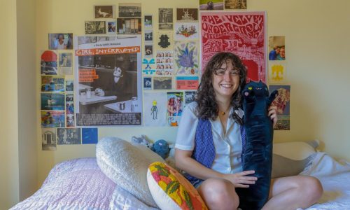 Wearing a white dress shirt and blue checkered vest, Lizzie England poses for the camera while sitting on her bed. Behind the bed is a collage of posters and images.