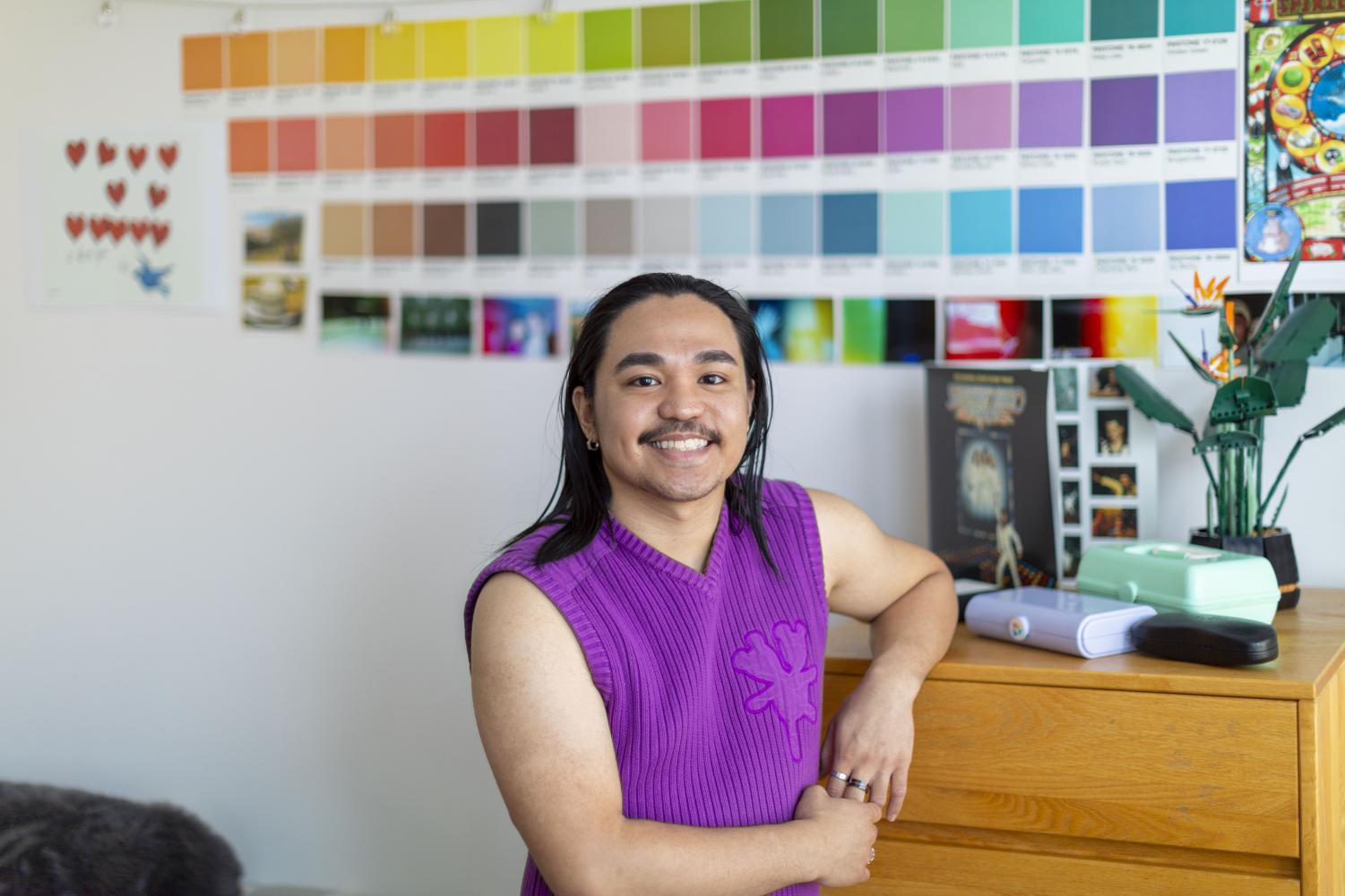 Wearing a sleeveless purple sweater, Khym Valenzuela looks at the camera with a smiling expression in his Gramercy dorm room. His left arm is resting on the top of his dresser. On the dresser, sits a green Lego plant. Khym is standing in front of his wall that has multi-colored pantone swatches.