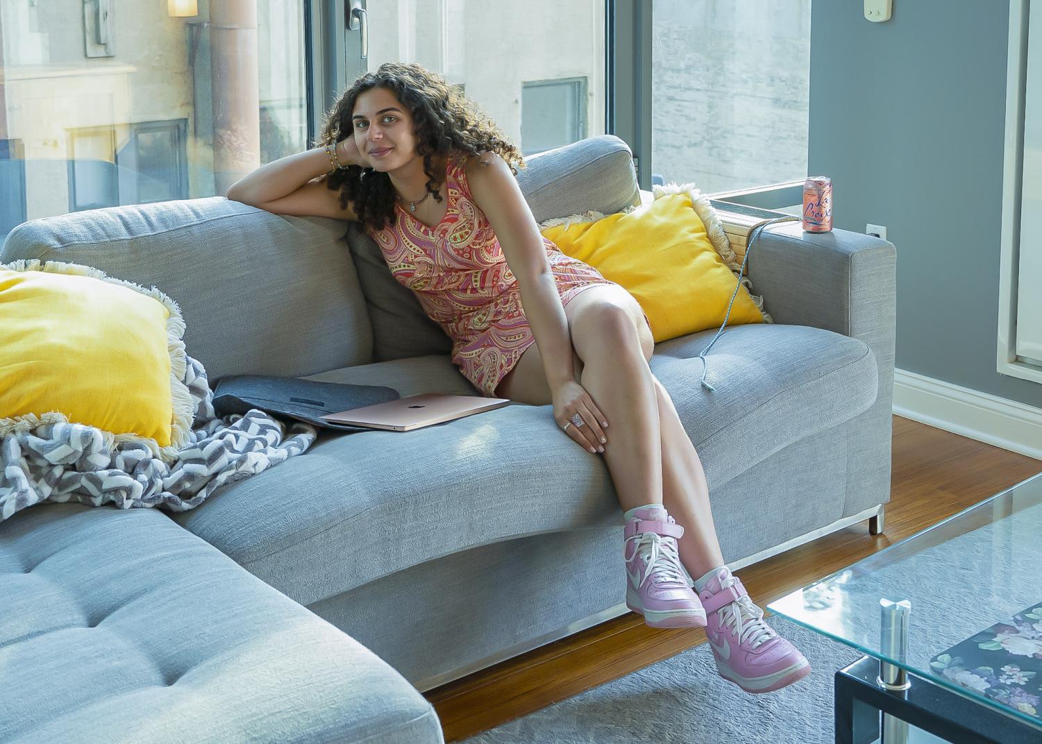 Adora Dayani in a pink-and-yellow patterned dress sits on a gray couch accompanied by yellow pillows.
