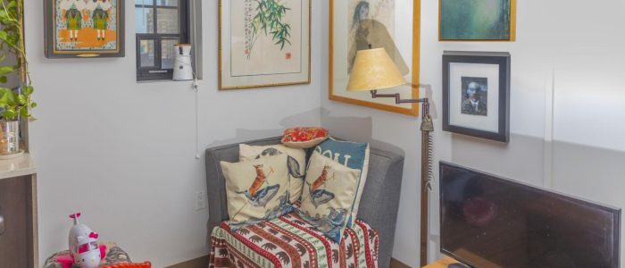The corner of John Moran and Kenneth French’s living room features a gray sitting chair with four pillows next to an ornate floor lamp and a small TV. The walls feature various pieces of artwork.