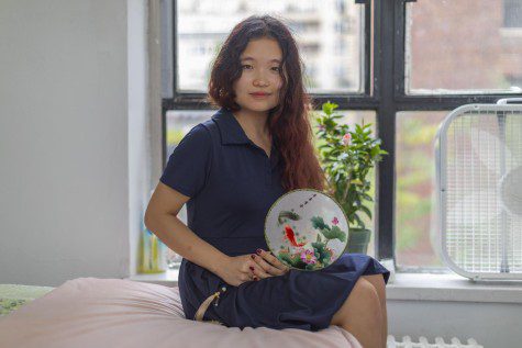 Sophia Wang looks at the camera with a pleasant expression. Sophia is wearing a dark blue collared dress and is holding a circular transparent object adorned with designs containing fish and flowers.