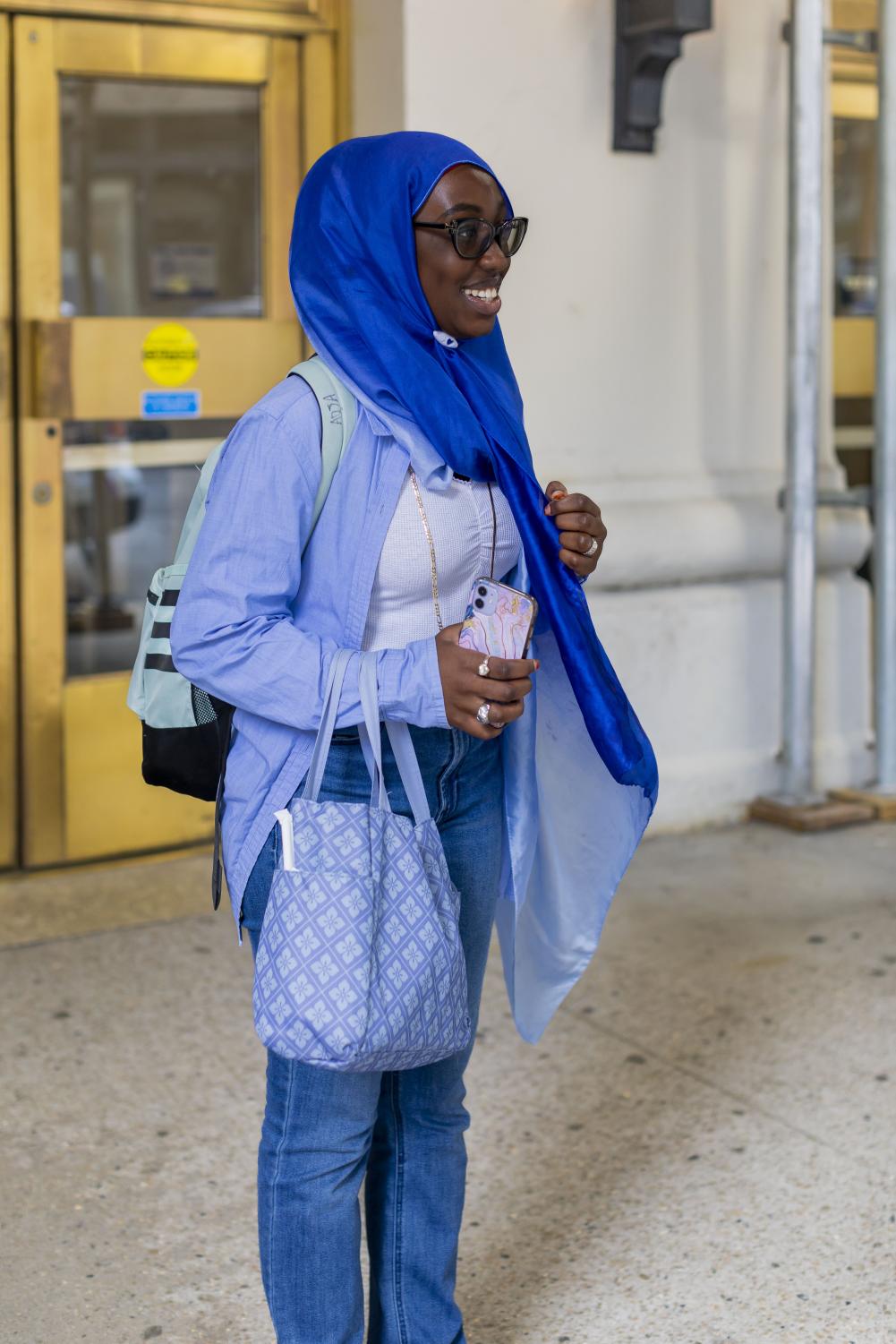 Aeja Seck stands outside of a College of Arts and Science building wearing jeans, a white shirt, a blue unbuttoned shirt and a head covering.