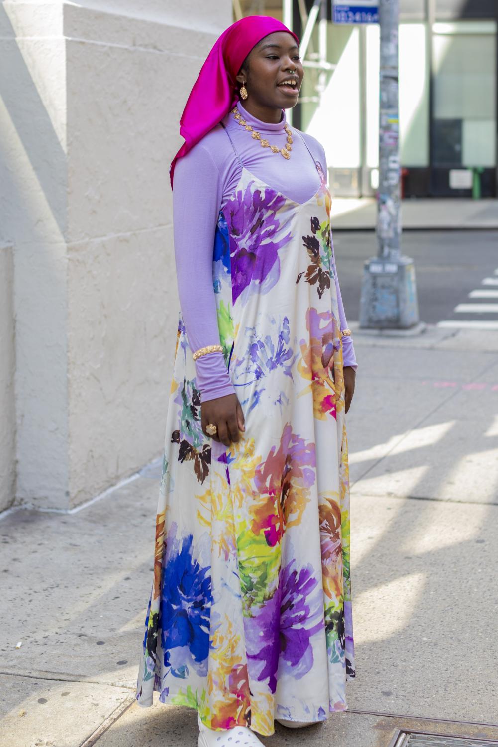 Hairiya Maiyaki is standing outside the College of Arts and Science wearing a floral maxi dress over a lilac long-sleeve shirt. Maiyaki wears a bright pink satin head covering.
