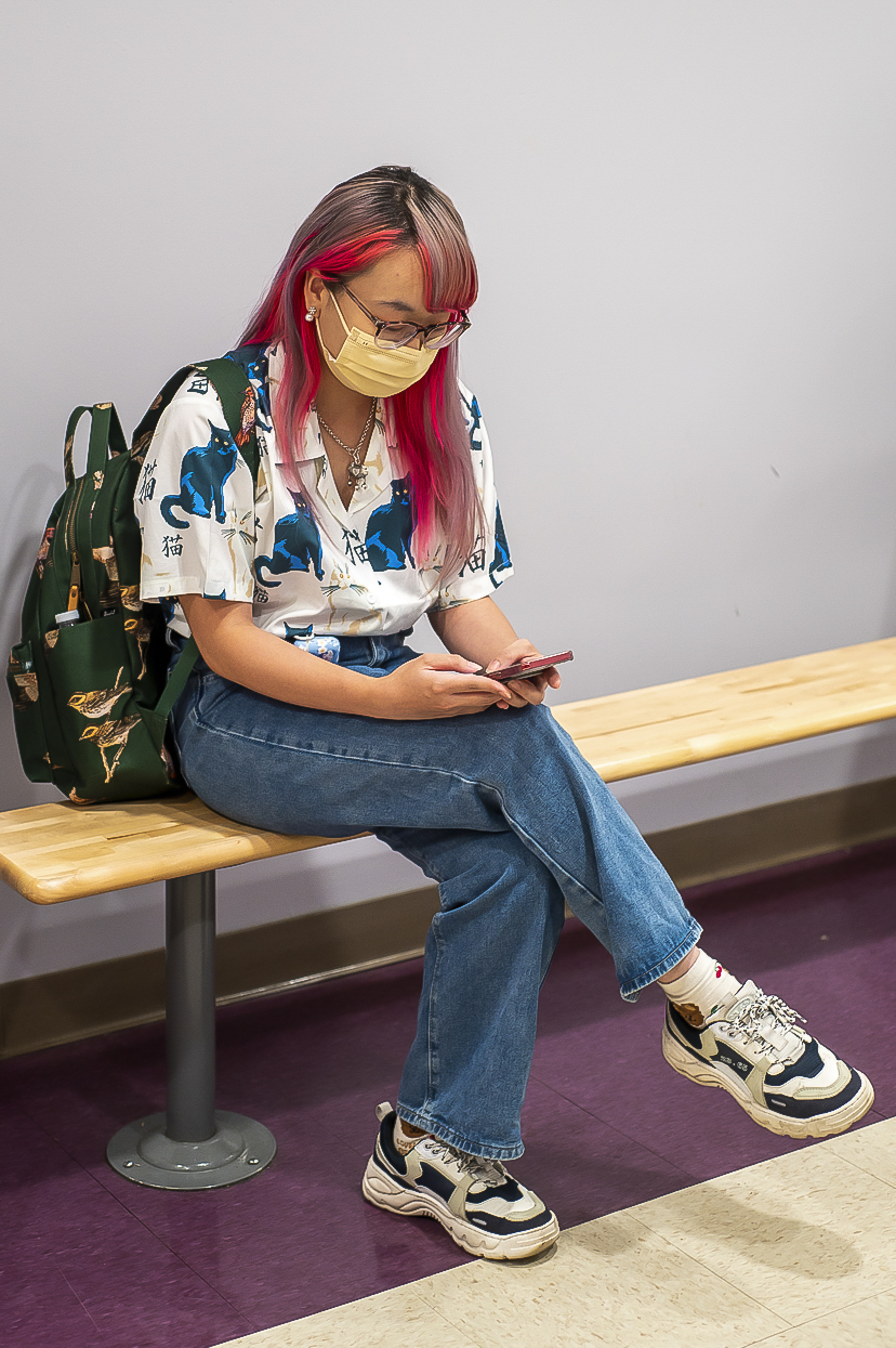 Mian Hua Zheng sits inside of a College of Arts and Science building looking at a phone and wearing jeans and a patterned shirt.