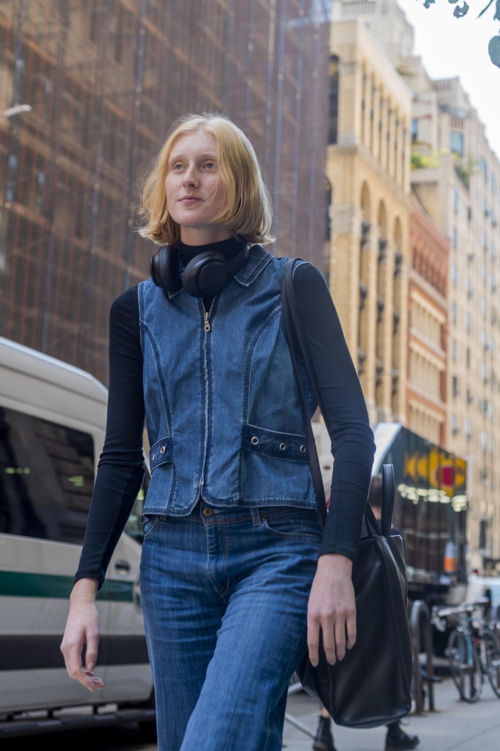 Hannah Wheless is walking down the street wearing blue denim pants and a zip-up denim vest over a black long-sleeve shirt.