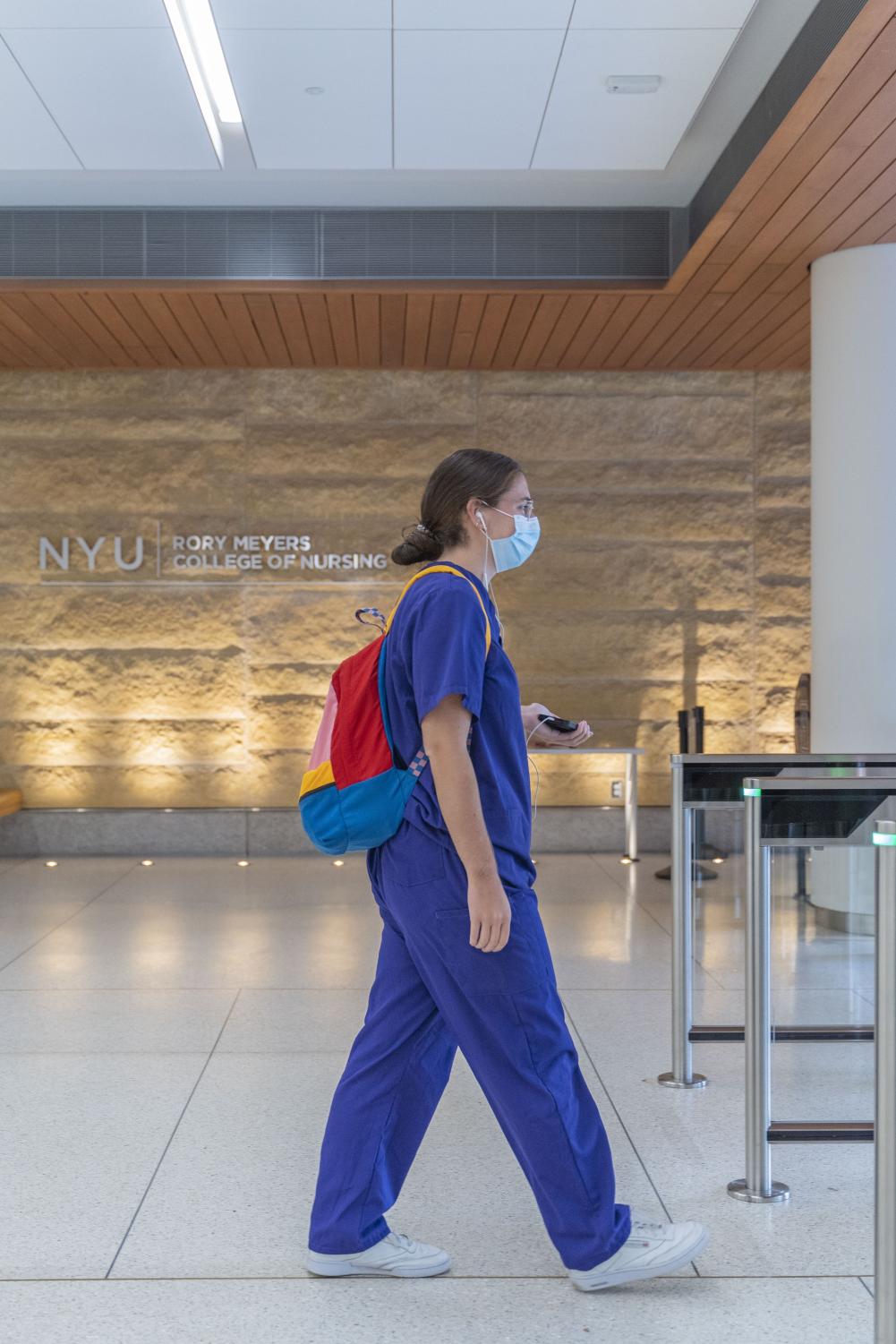 Jessica Alvarez walks in the lobby of a Meyers building wearing purple scrubs and a multicolored backpack.