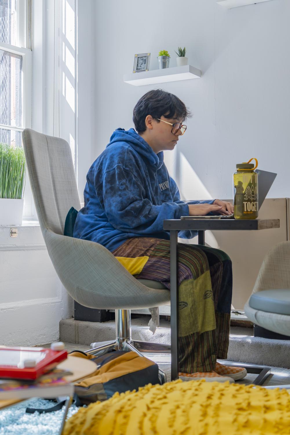 Virgile-Minh Perrier works on a computer while sitting in a Silver building. Perrier is wearing a blue tie-dye sweatshirt and multi-pattern patchwork pants.