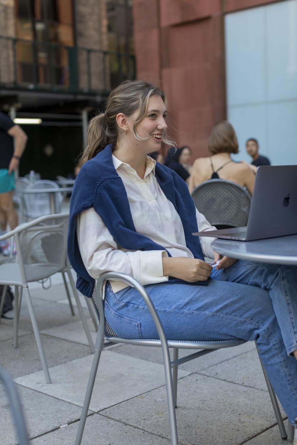 Isabella Perrigo sits at a table outside of a Stern building while talking to a friend. Perrigo is wearing a white button-up shirt, a blue cardigan draped over her shoulders and blue jeans.
