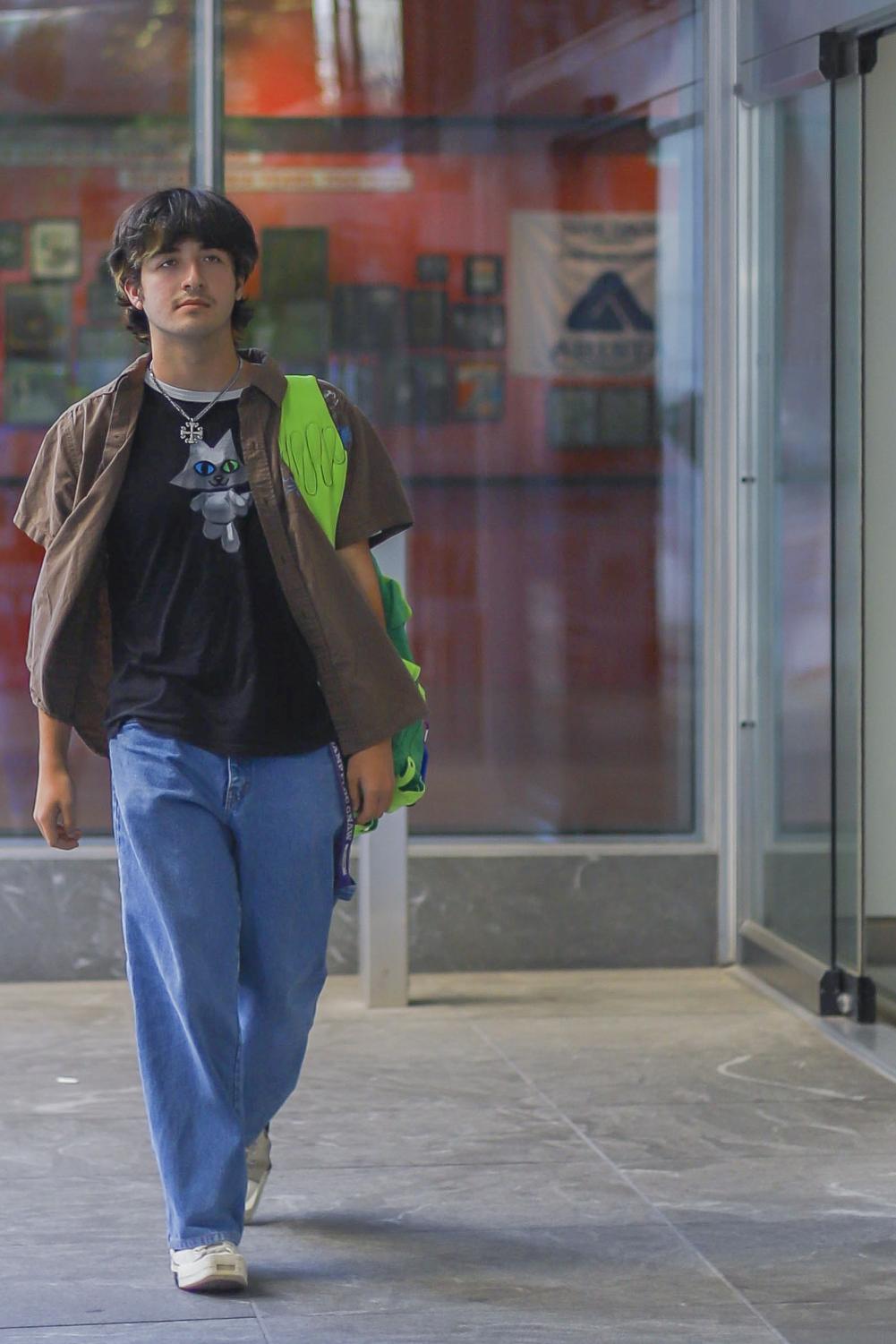 Matthew Toups walks outside of a Tandon building wearing a black graphic t-shirt, brown dress shirt, baggy blue jeans and a green backpack.