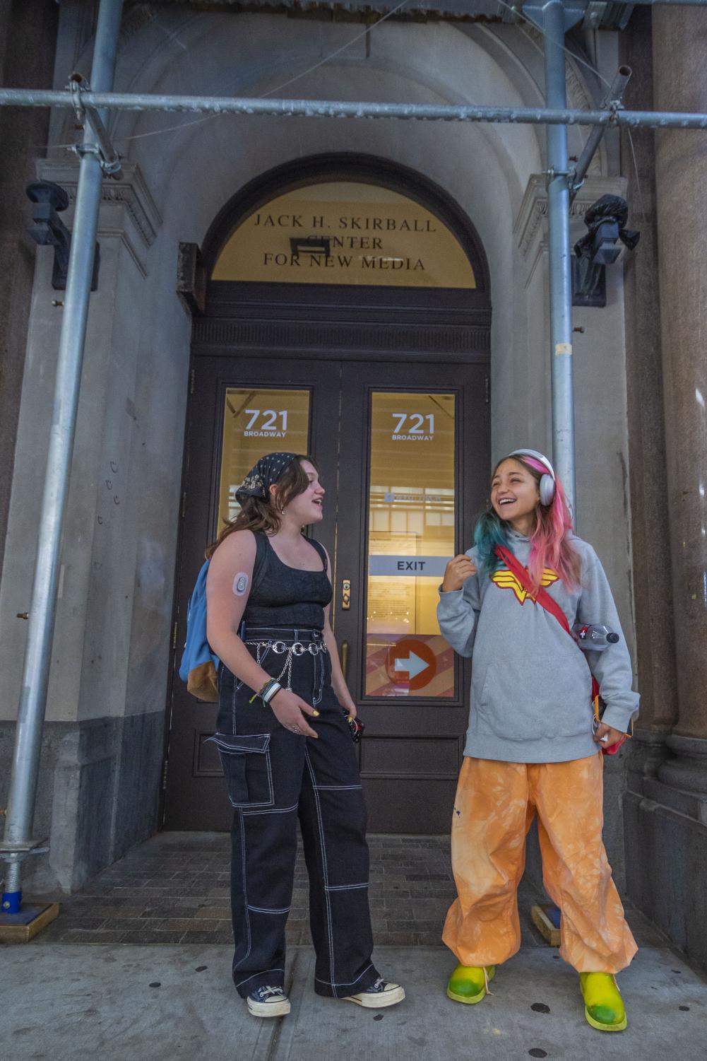 Cassie Lustig (left) and Moriah Reibman (right) are laughing as they stand outside of a Tisch building. Cassie is wearing a black tank top, black denim pants with white stitching, a blue backpack and a chain belt. Moriah is wearing a graphic gray hoodie, orange baggy cargo pants and a red bag.