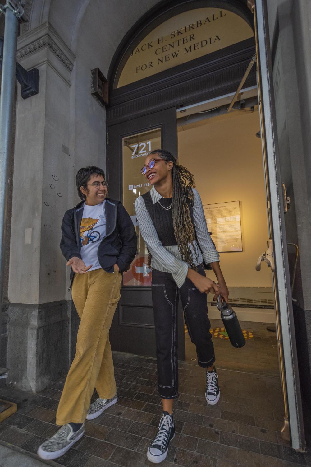 Janelle Hernandez (left) and Niya Leigh (right) smile at one another as they walk out of a Tisch building. Janelle is wearing a white graphic t-shirt, a black windbreaker and muted yellow corduroy pants. Niya is wearing a vertically striped gray dress shirt, black cropped cable-knit sweater and black denim pants with white stitching.