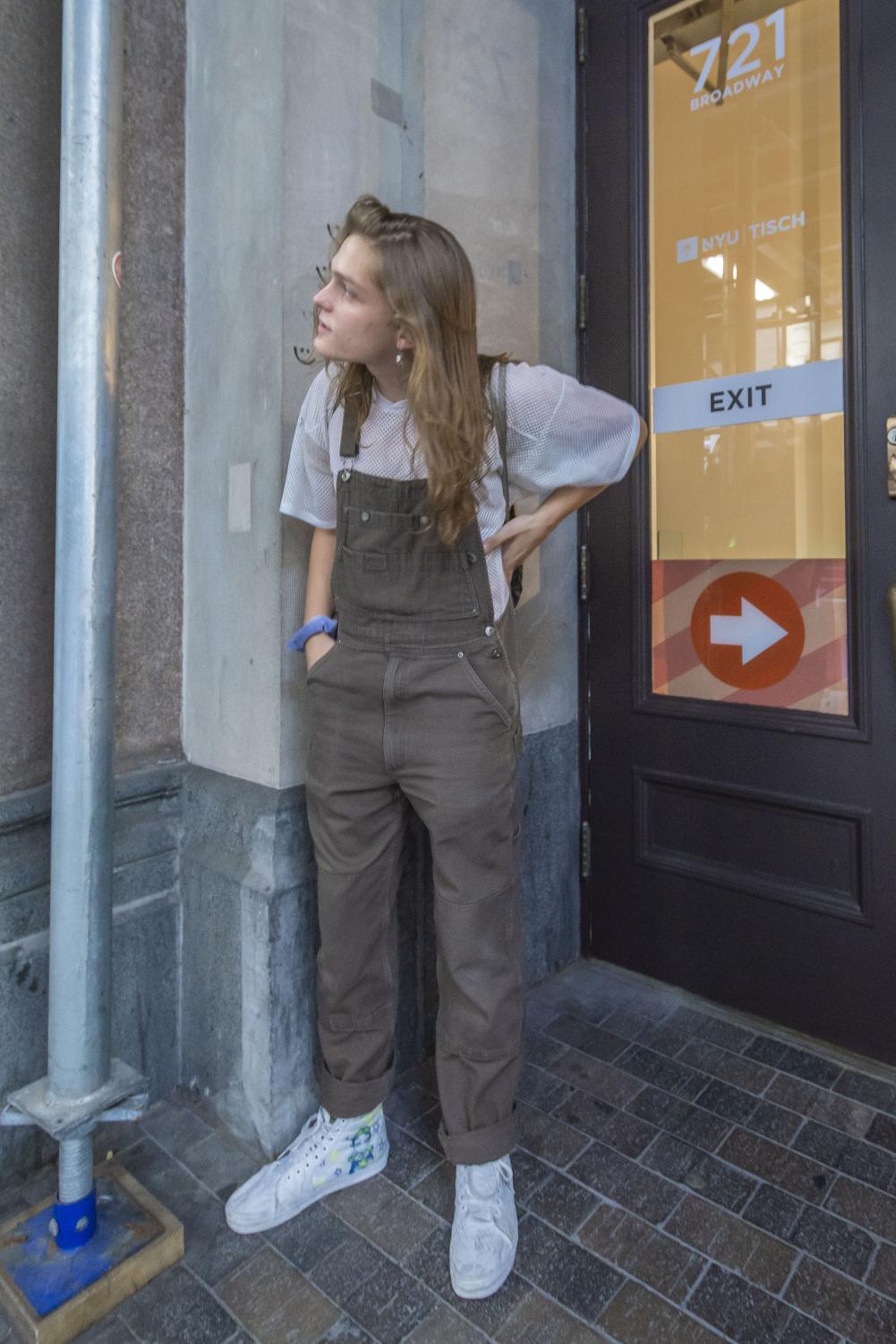 Rowan Trowbridge-Wheeler stands in front of a Tisch building looking away from the camera. Trowbridge-Wheeler is wearing a white mesh t-shirt with brown overalls.