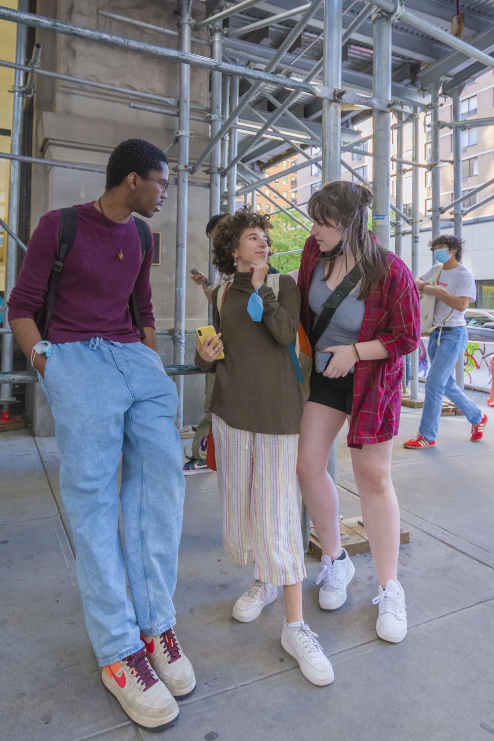 Ahmad Goode (left), Michela Richards (middle) and Blake Chrusciel (right) have a conversation while leaning on scaffolding in front of Tisch. Ahmad is wearing a dark magenta sweater and baggy blue jeans. Michela is wearing an olive turtleneck sweater and linen pants with vertical pastel stripes. Blake is wearing a gray tank top, red plaid dress shirt and black shorts.