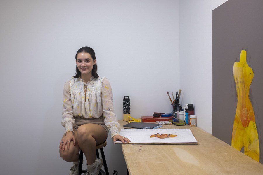 Ella Zona, dressed in a white blouse, sits in her studio with white walls. To the camera right of Zona, her desk is filled with her tools and with two unfinished paintings on top.