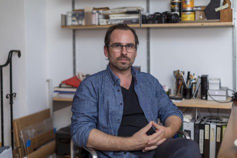 Professor Anthony Graves sits in front of his desk in his studio wearing glasses and dressed in a blue denim jacket and black shirt.