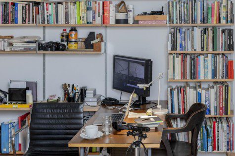 Professor Anthony Grave’s studio is filled with a table, two chairs and a shelf. Atop of the table is a computer setup. Atop of the shelf are digital cameras and a collection of books.