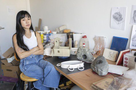 Lizzy Chemel, dressed in a white tank top and a pair of blue denim jeans, sits in her studio.